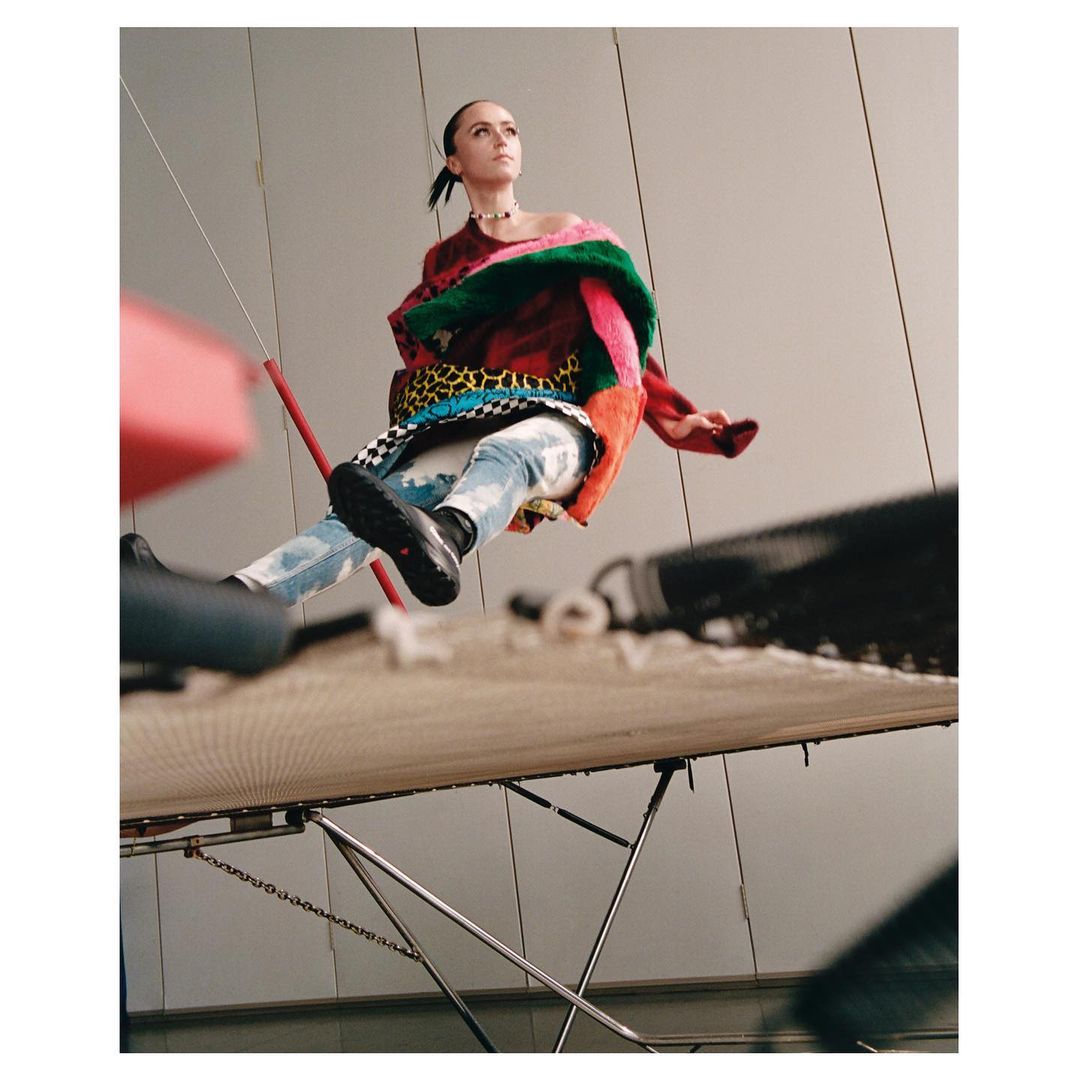 Fashion editorial shot from a low angle captures a woman in an oversized multicolored sweater, light-wash jeans, and black shoes.