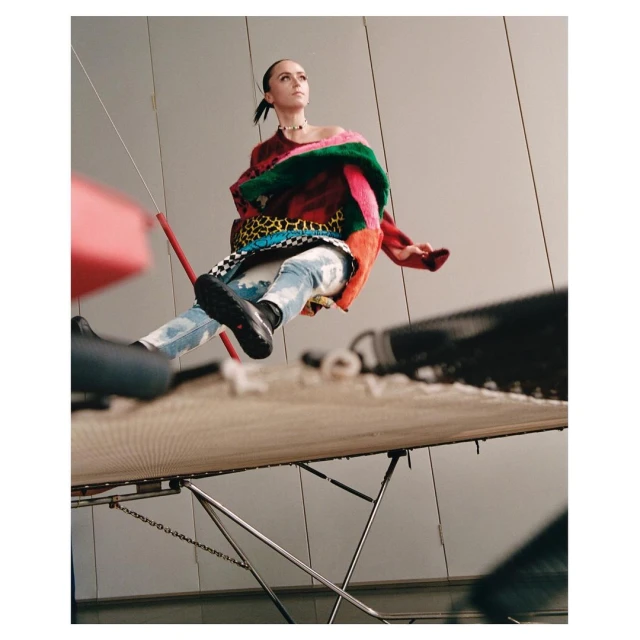 Fashion editorial shot from a low angle captures a woman in an oversized multicolored sweater, light-wash jeans, and black shoes.