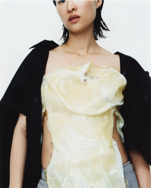 Fashion portrait of an Asian woman in a translucent top and black jacket against a white backdrop, showcasing contemporary style.