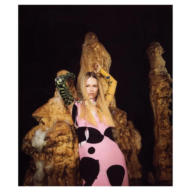 Fashionable woman with long blonde hair poses in a pink and black dress with patterned sleeves against a backdrop of rock formations.