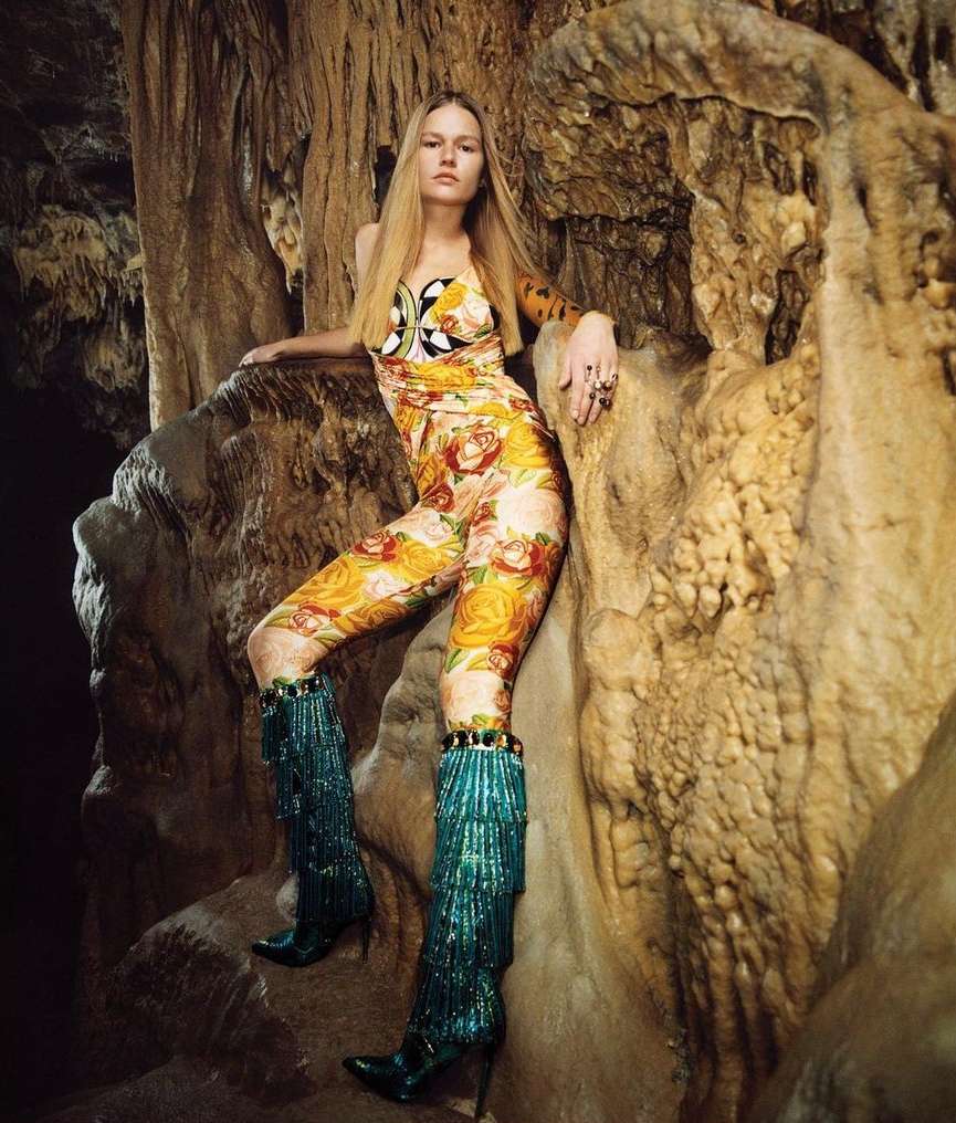 Woman in a floral jumpsuit and teal fringe boots poses inside a cave with tan rock formations and dim lighting, fashion editorial style.