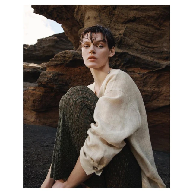 Woman sits on a black sand beach near a rocky cliff. She wears a linen shirt and textured pants in an outdoor fashion shot.