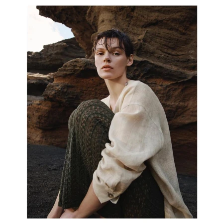 Woman sits on a black sand beach near a rocky cliff. She wears a linen shirt and textured pants in an outdoor fashion shot.