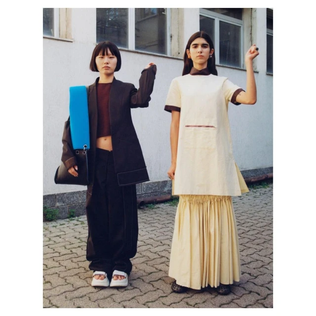 Two young women in fashionable neutral-toned outfits stand outdoors in front of a building, one with her fist raised in the air.