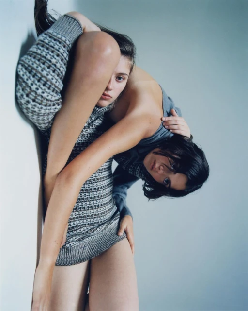 Two women in a studio setting. One woman stands tall wearing a patterned dress, while the other is positioned horizontally behind her.