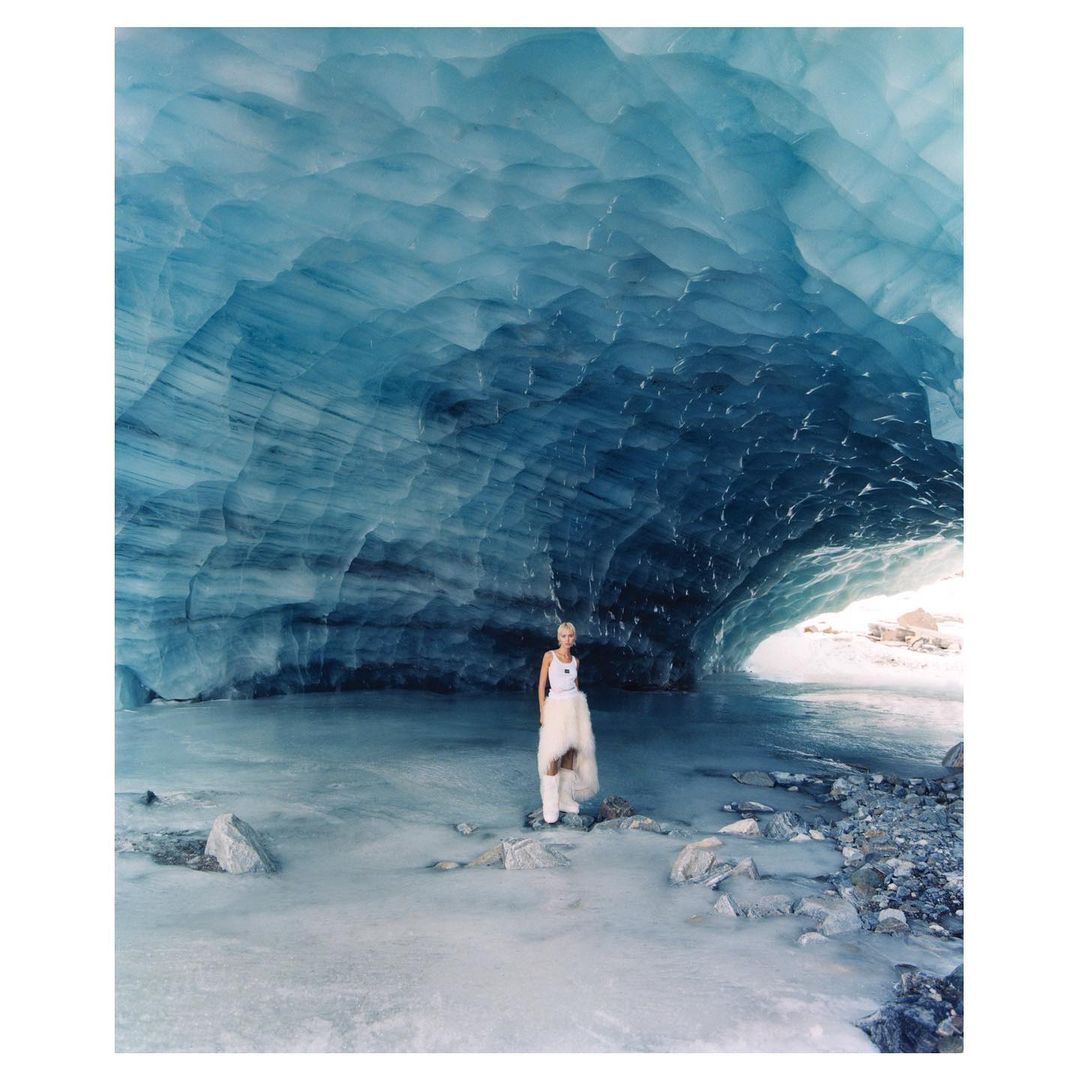Woman standing in a glacial ice cave with blue ice formations and a stream of water, wearing a white top and light-colored skirt.