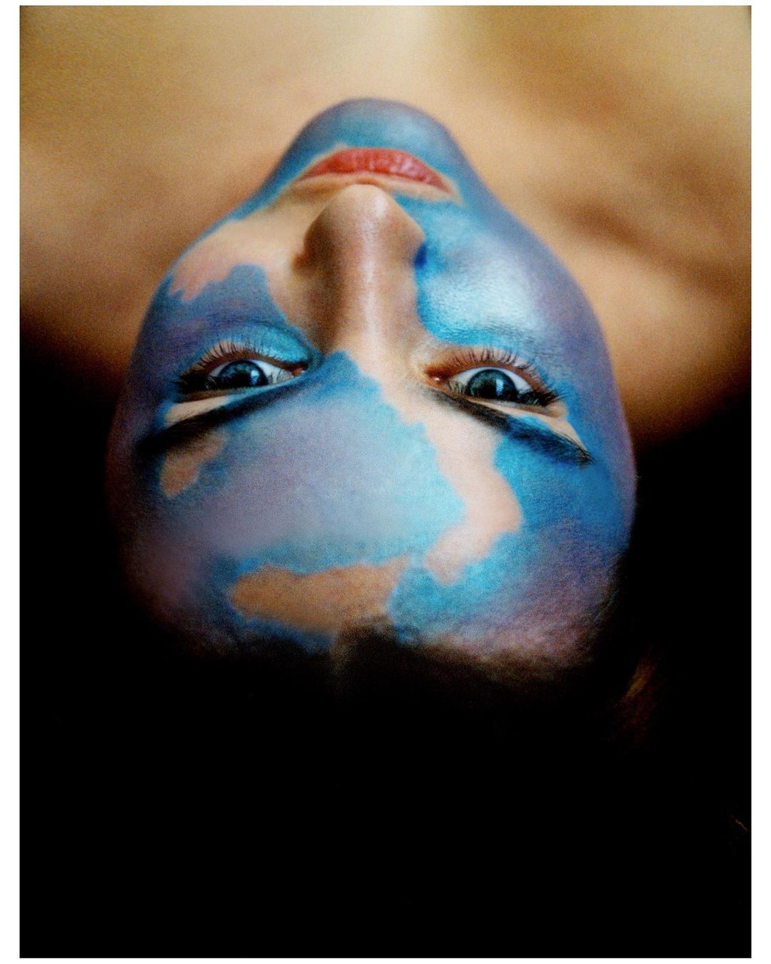 Close-up portrait of a person with artistic blue face paint and red lips, photographed upside down by Dario Catellani.