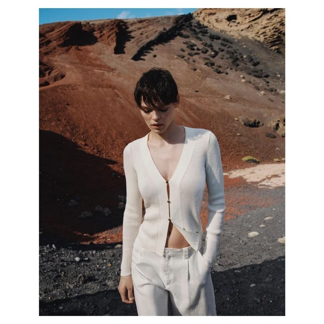 Woman in white cardigan and pants poses in a barren landscape with a textured mountain backdrop under natural lighting.
