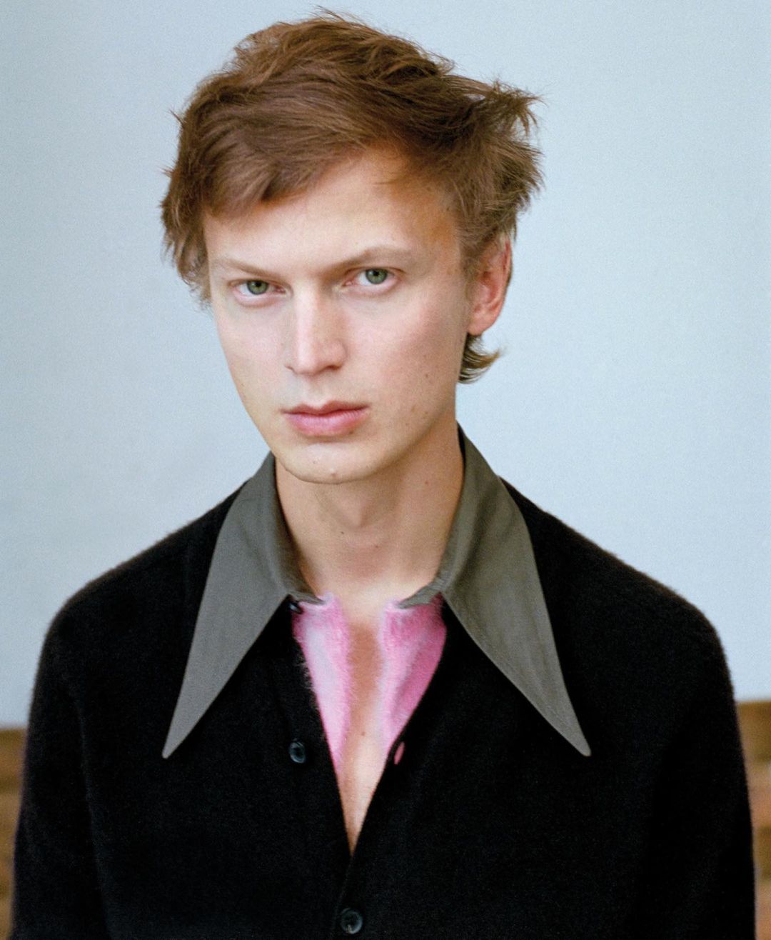 Portrait of a young adult male with reddish-brown hair wearing a black cardigan and gray collar shirt against a light backdrop.