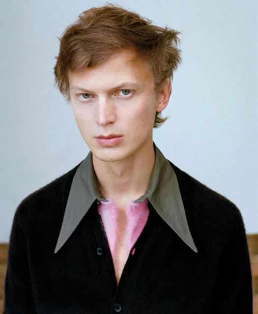 Portrait of a young adult male with reddish-brown hair wearing a black cardigan and gray collar shirt against a light backdrop.