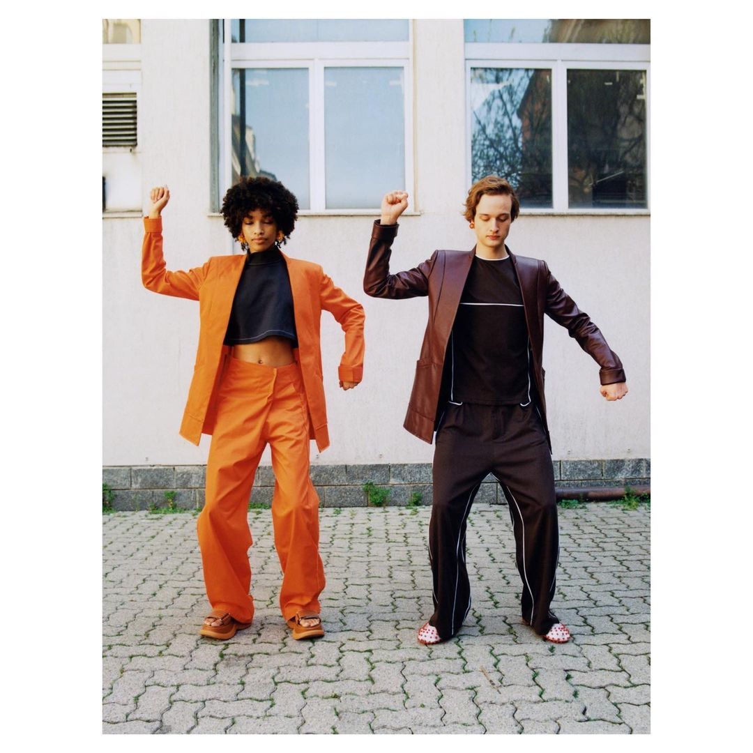 Two people in fashionable 1990s style clothing strike a pose outdoors. A woman in orange and a man in brown stand on cobblestones.
