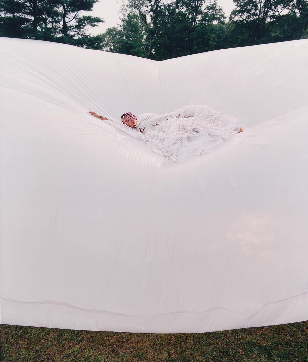 An adult woman lying on a large, white, inflated surface in an outdoor setting with trees visible in the background, wearing a lacy white gown.
