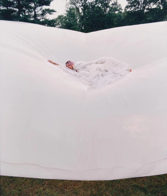 An adult woman lying on a large, white, inflated surface in an outdoor setting with trees visible in the background, wearing a lacy white gown.