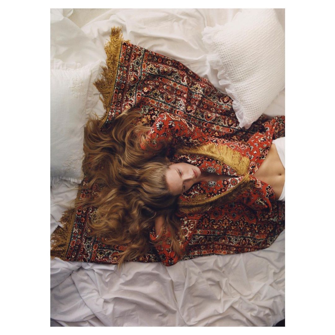 Woman lying on white bed covered by patterned textile. Long brown hair spread across a bedsheet with soft pillows and a relaxed pose.