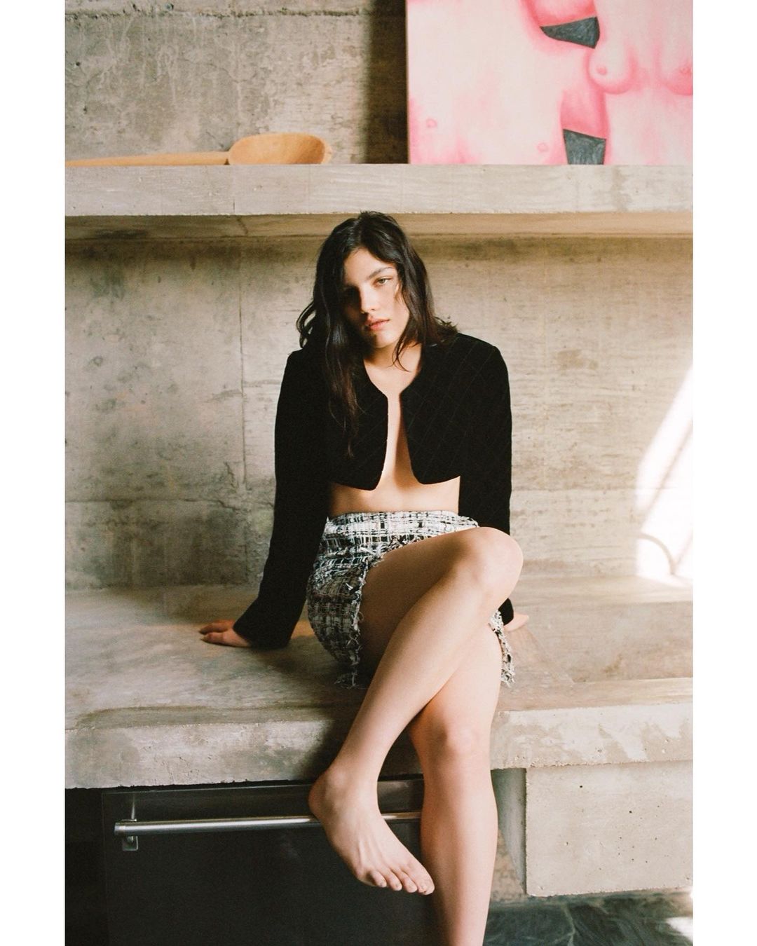 A woman poses in a black jacket and tweed skirt while seated against a concrete wall, accented by a wooden spoon and abstract painting.