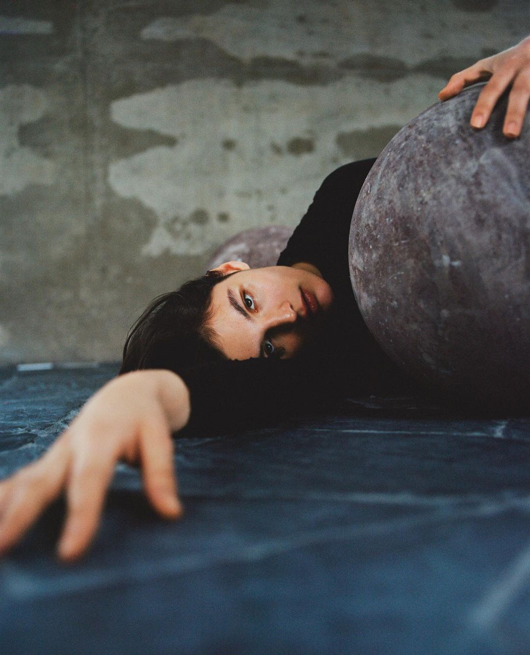Woman with dark hair poses indoors with large grey spheres in a dimly lit room, with her arm outstretched and dark clothing.