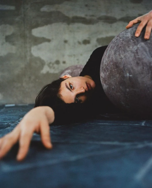 Woman with dark hair poses indoors with large grey spheres in a dimly lit room, with her arm outstretched and dark clothing.