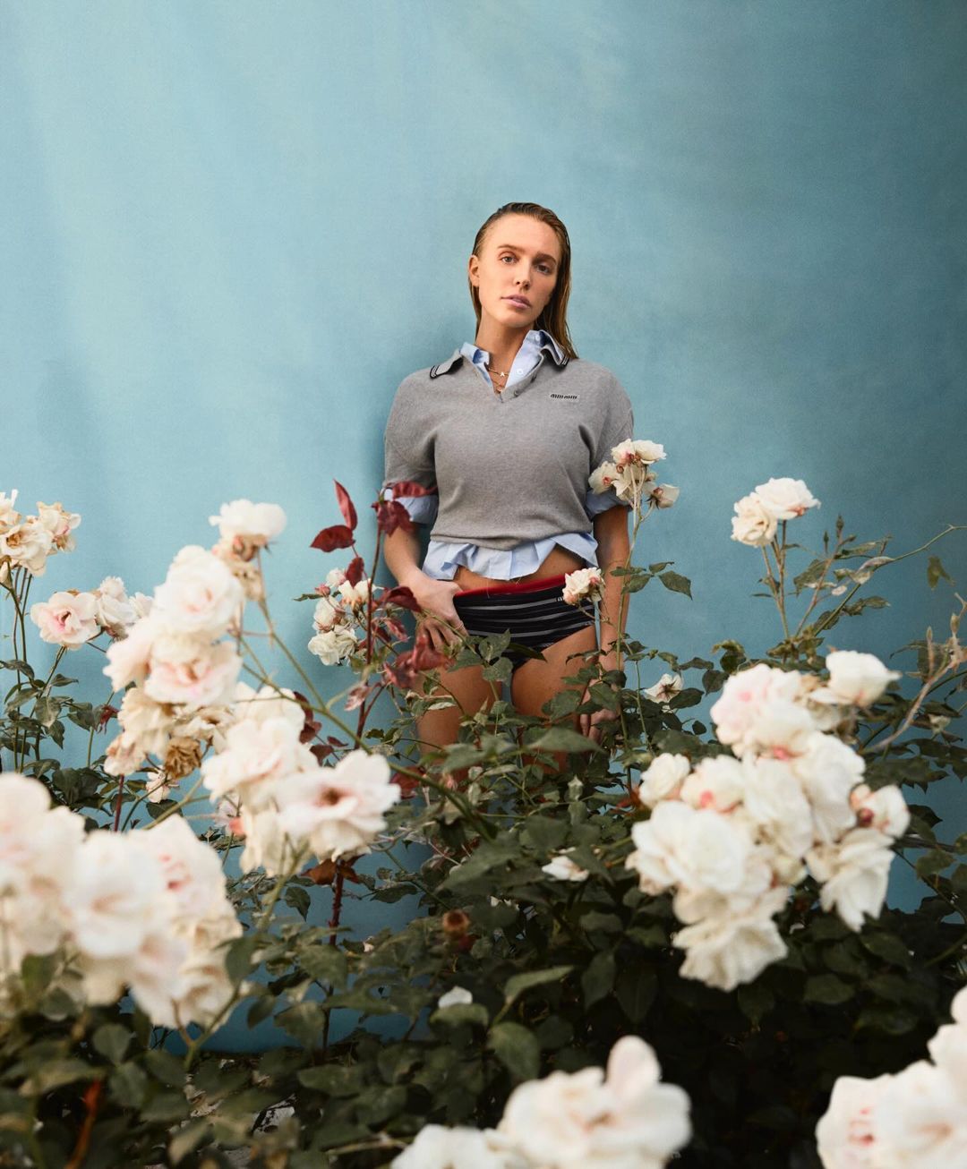 A woman stands in front of a blue wall surrounded by white roses, wearing a grey sweater, blue button-down shirt, and striped bottoms.