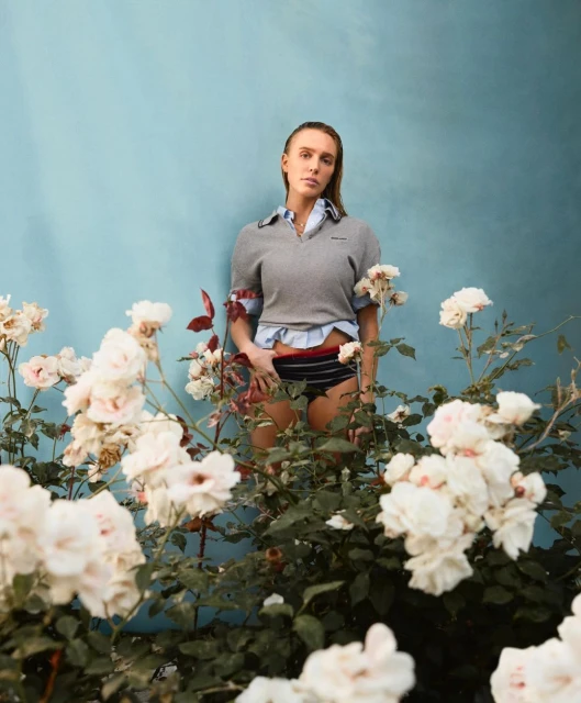 A woman stands in front of a blue wall surrounded by white roses, wearing a grey sweater, blue button-down shirt, and striped bottoms.