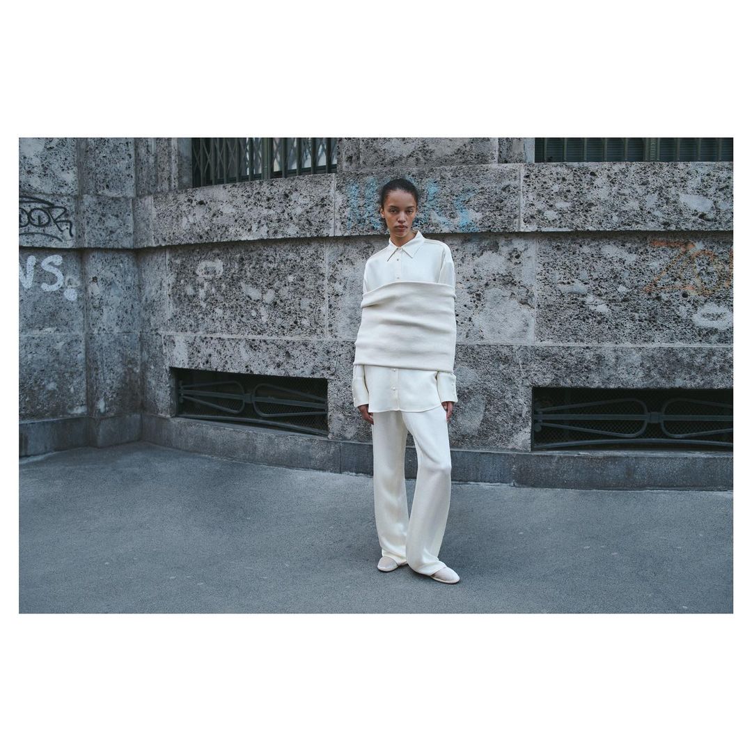 Fashion shot of a woman outdoors wearing an oversized white shirt and wide-leg trousers, standing against a textured wall in daylight.