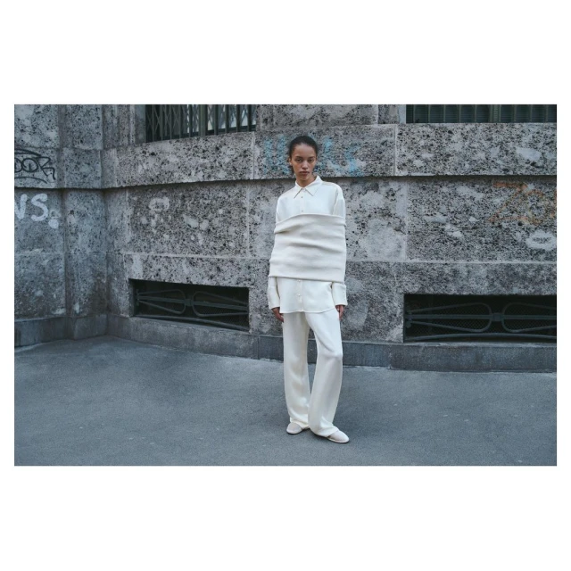 Fashion shot of a woman outdoors wearing an oversized white shirt and wide-leg trousers, standing against a textured wall in daylight.