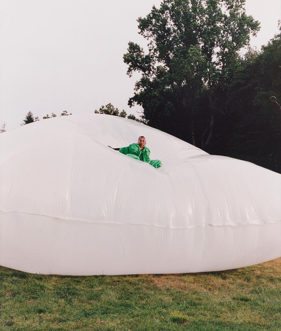 Adult in green jumpsuit reclines inside large, inflated white plastic on grassy field with trees in background on overcast day.