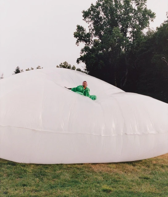 Adult in green jumpsuit reclines inside large, inflated white plastic on grassy field with trees in background on overcast day.