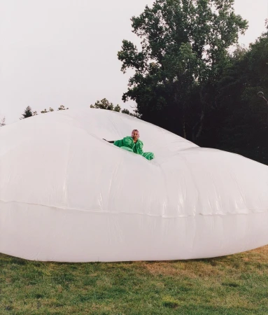 Adult in green jumpsuit reclines inside large, inflated white plastic on grassy field with trees in background on overcast day.