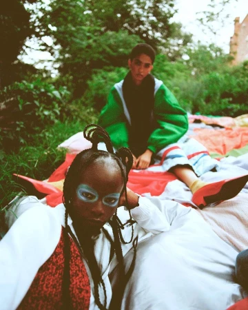 Two models outdoors on a patterned blanket, one in the foreground with blue eye makeup and braided hair, another seated behind wearing a green jacket.