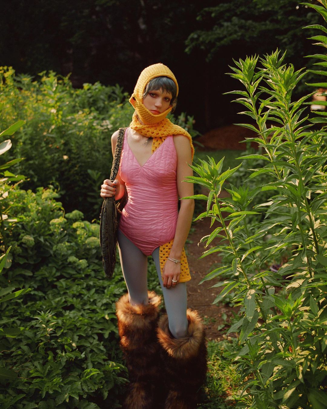 A woman in a pink swimsuit and fur boots stands on a path surrounded by lush greenery in a fashion editorial-style shot.