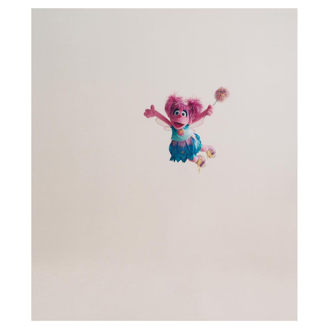 Abby Cadabby, the pink Muppet fairy from Sesame Street, is seen in a full shot against a pale background holding a magic wand.