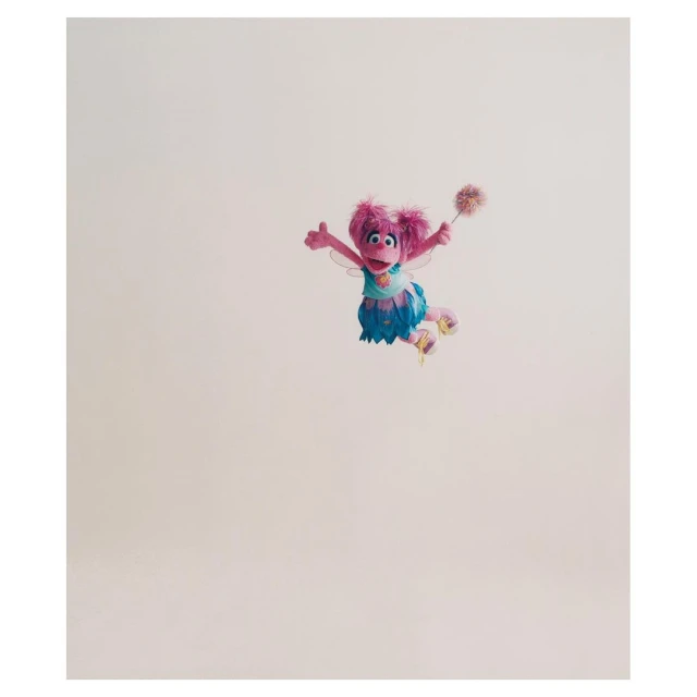 Abby Cadabby, the pink Muppet fairy from Sesame Street, is seen in a full shot against a pale background holding a magic wand.