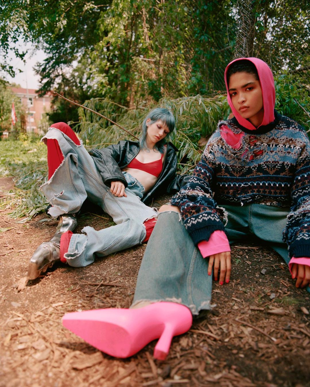Two women in outdoor fashion shoot, one in pink hoodie and patterned sweater, the other in a leather jacket and distressed jeans, posed in forest.