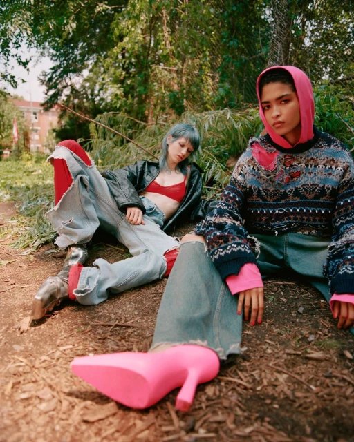 Two women in outdoor fashion shoot, one in pink hoodie and patterned sweater, the other in a leather jacket and distressed jeans, posed in forest.