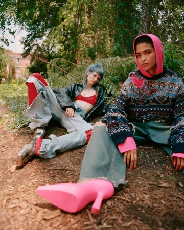 Two women in outdoor fashion shoot, one in pink hoodie and patterned sweater, the other in a leather jacket and distressed jeans, posed in forest.