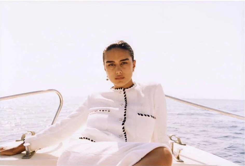 Elegant adult female model poses on a boat wearing a cream-colored dress with black trim on sunny day by a blue water.