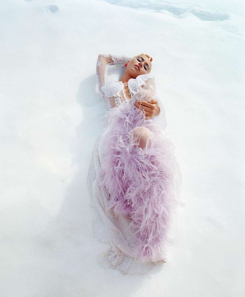 Adult female model lies on snow in an off-white dress with lavender feathered boa. She has fair skin, short light hair, and dark eye makeup.