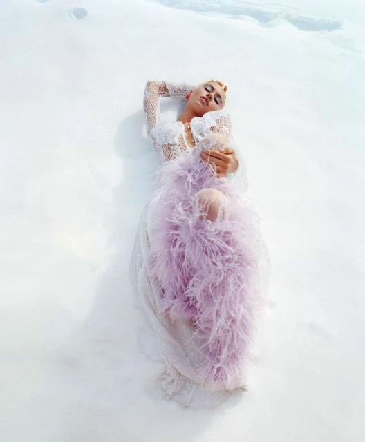Adult female model lies on snow in an off-white dress with lavender feathered boa. She has fair skin, short light hair, and dark eye makeup.