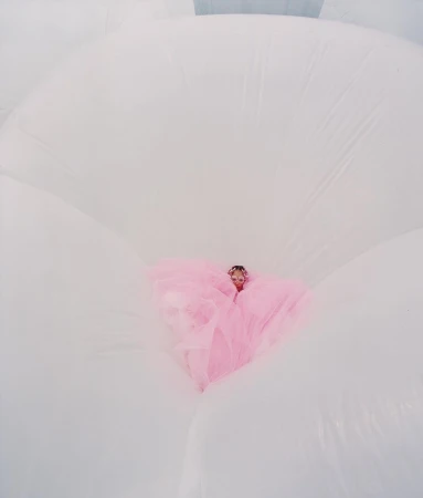 Woman in a pink dress surrounded by large, white inflatable shapes, creating a dreamy, ethereal aesthetic with a unique perspective.