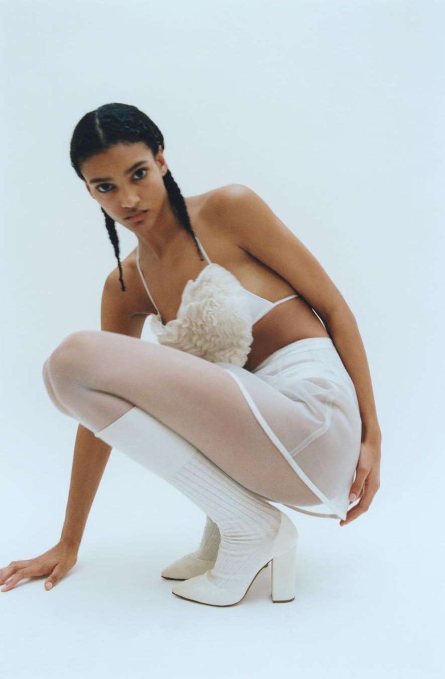 A young woman with braids, wearing a white top and skirt with knee-high boots, is squatting in a photo studio with a light background.