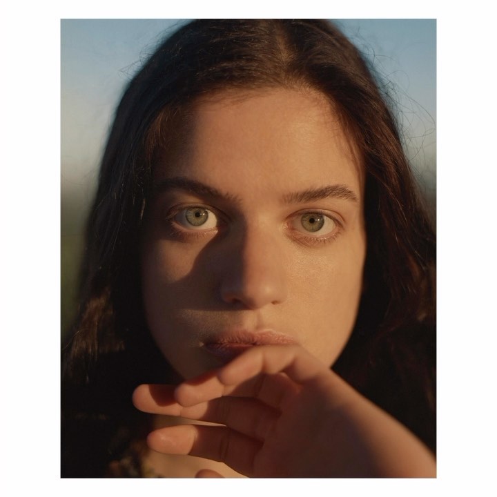 Close-up portrait of a young adult female with long brown hair and light green eyes, softly illuminated in outdoor setting.
