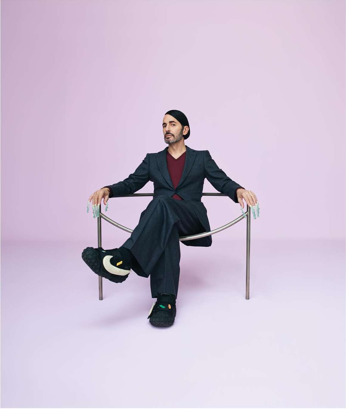 Stylish adult man in a dark suit and knit cap, sits in a metal chair against a lilac background in a studio setting.