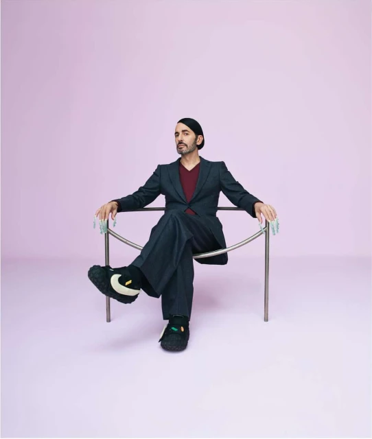 Stylish adult man in a dark suit and knit cap, sits in a metal chair against a lilac background in a studio setting.  