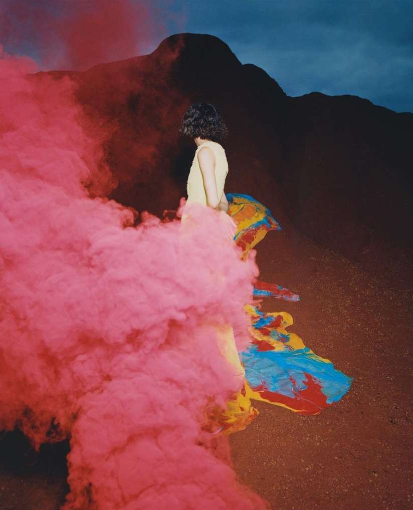 An adult female stands in a desert landscape next to pink smoke wearing a pastel yellow dress and a draped blue and yellow cloth.