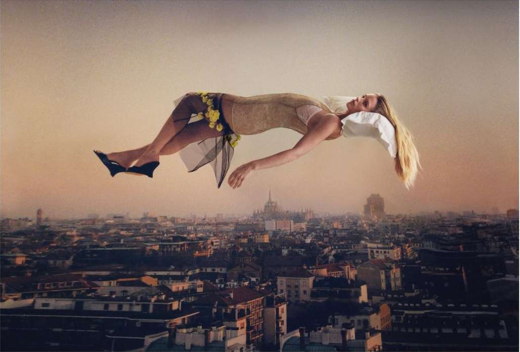 Surreal image of a blonde woman floating in mid-air with a pillow beneath her head, above a cityscape at dusk, wearing heels and a dress.