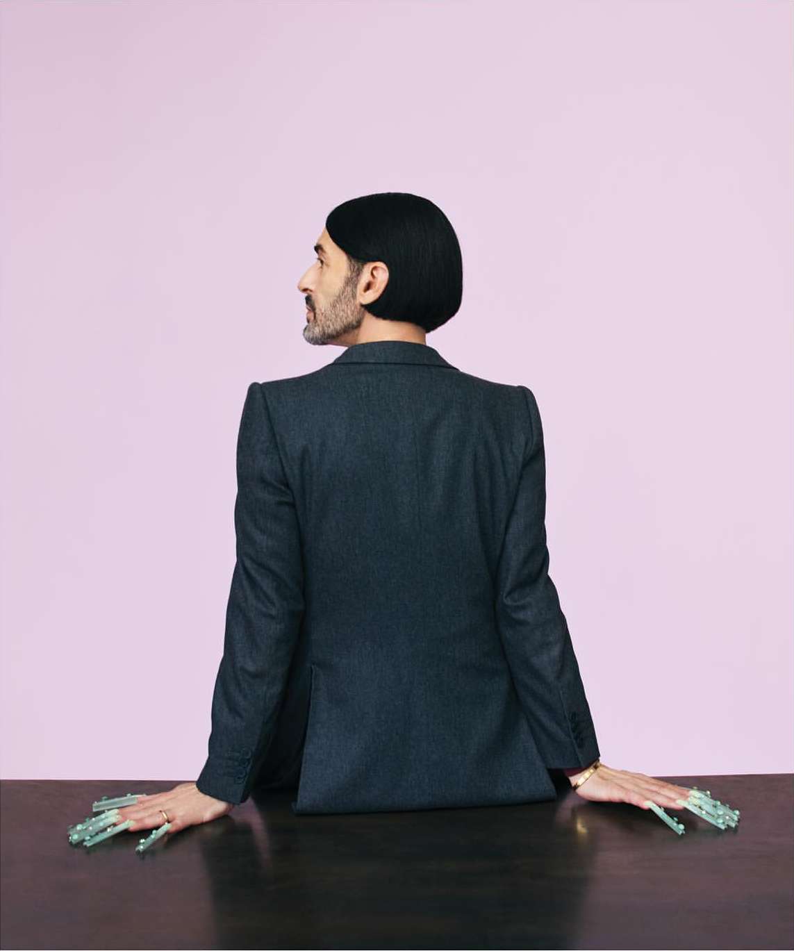 Adult male in suit with long decorative nails poses behind a wooden table against a lilac background in a studio shot.