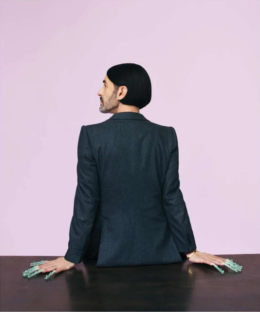 Adult male in suit with long decorative nails poses behind a wooden table against a lilac background in a studio shot.