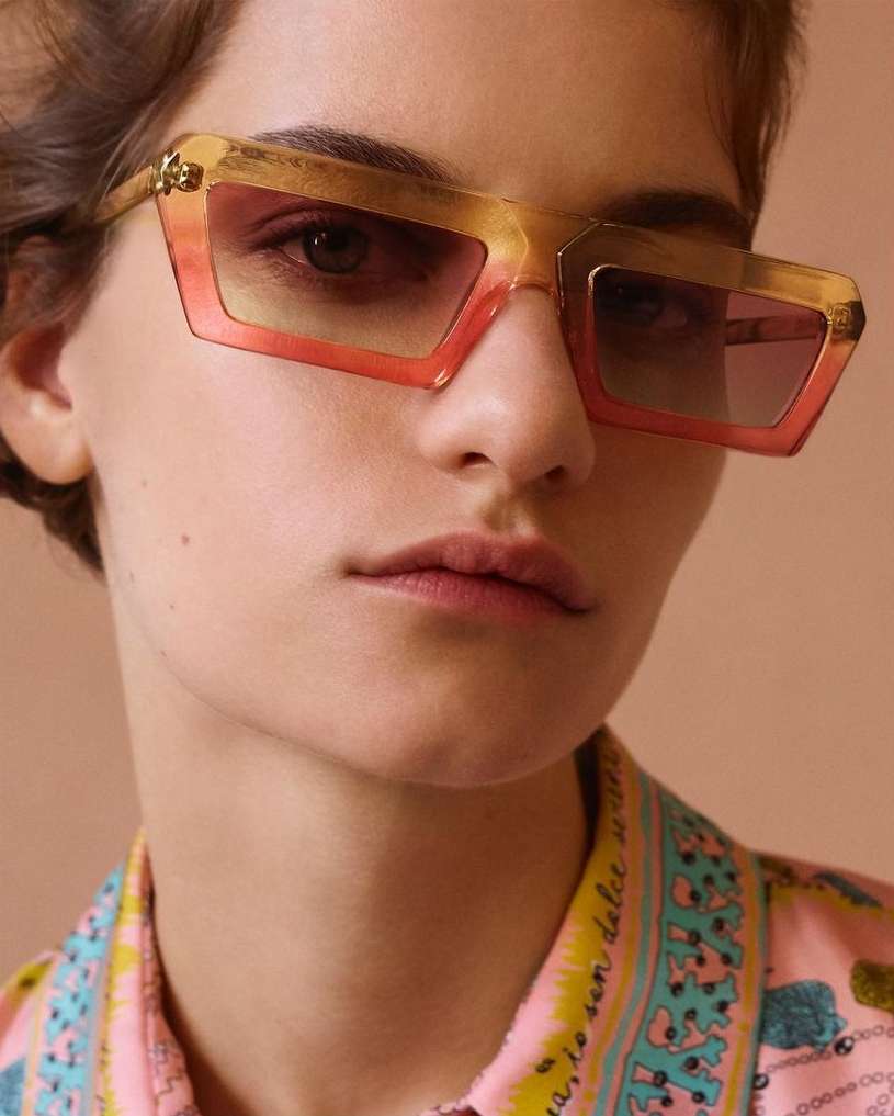 A young woman with fair skin, wearing stylish orange sunglasses and a patterned pink shirt, poses in a close-up studio shot.