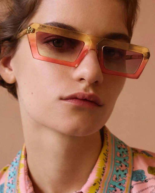 A young woman with fair skin, wearing stylish orange sunglasses and a patterned pink shirt, poses in a close-up studio shot.