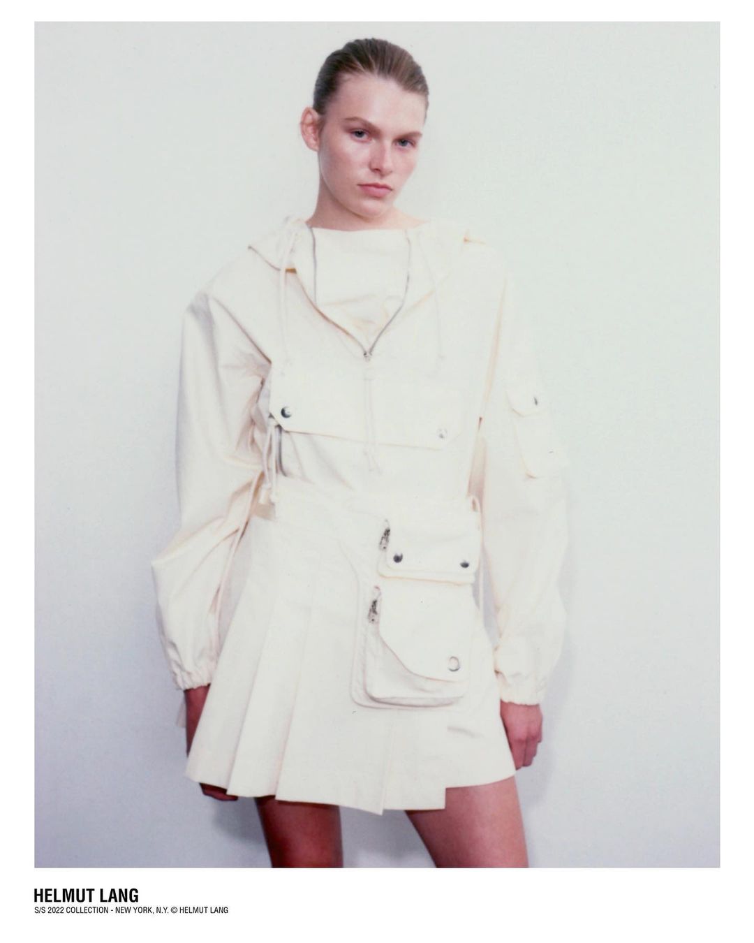 Model wearing a cream-colored utility jacket and pleated miniskirt against a plain light wall in a portrait by Dario Catellani.